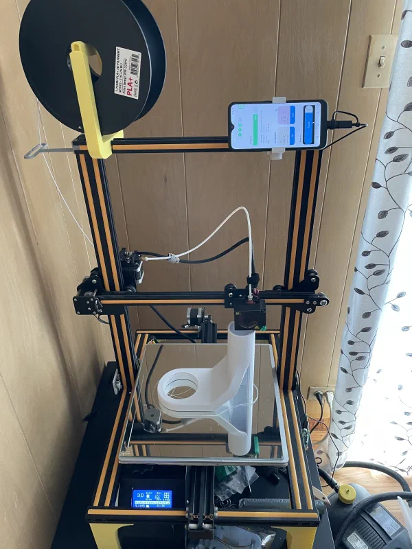Metal frame of a 3d printer with spool of filament attached to it on top, printing a white piece
