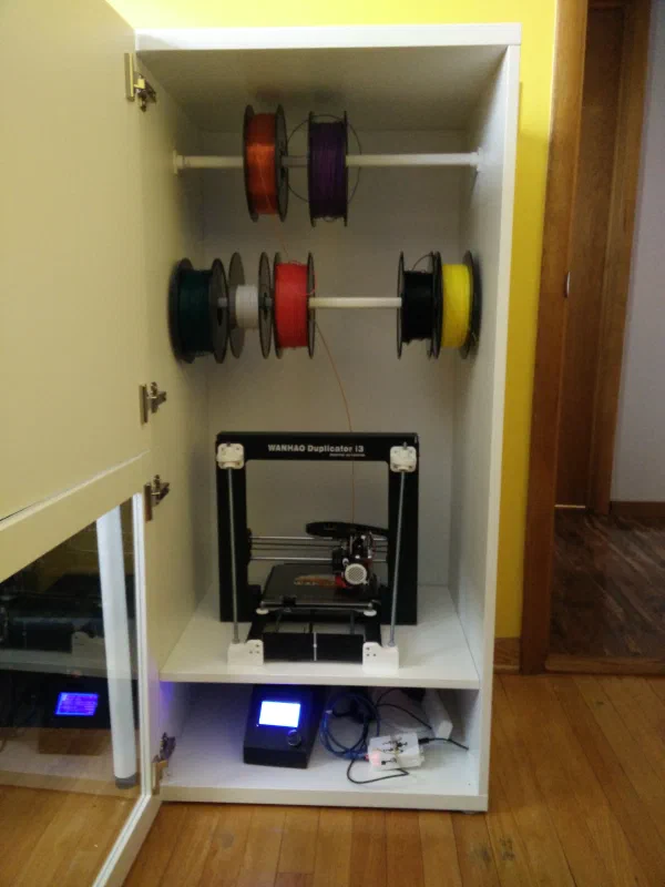 White cabinet with two shelves, top one containing a 3d printer with filament rolls hanging above, and bottom shelf containing electronics